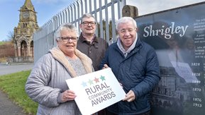 Three people hold a Rural Community Awards placard, beside a Shrigly sign.