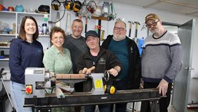 Six people stand by a lathe.