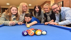 A youth prepares to play pool, watched by a small group of adults.