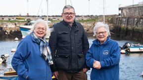 A man and two women with a small harbour behind them.