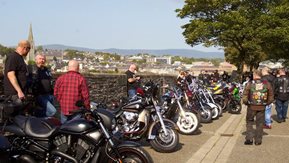 A number of men stand by their motorbikes by an old wall.