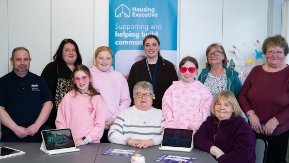 A group of people sit around a table smiling at the camera while using tablets. 