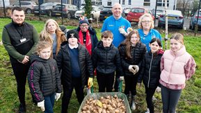 School-children and adults around a daffodil-filled wheelbarrow.