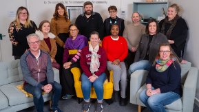 A group of men and women sit together on arm chairs smiling at the camera.  