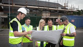 Six people in hi-vis jackets study plans on a building site.
