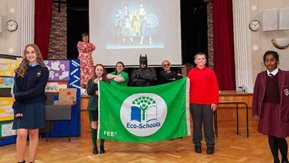 School children, adults and a man in a super hero costume appear with a large banner.