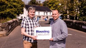 David Brownlee and Eoin McKinney pose with a Housing Executive sign on the bridge in Garrison in Fermanagh