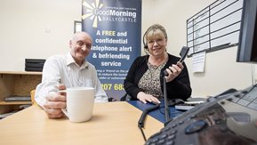 A man and woman sit at a desk. The woman holds a phone.