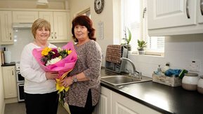 Two women stand in a kitchen with a bunch of flowers 